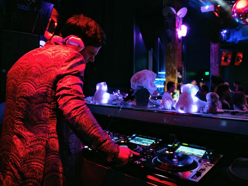 chillbill in a red brocade jacket mixing at Great Northern, white teddy bears lit up along the booth