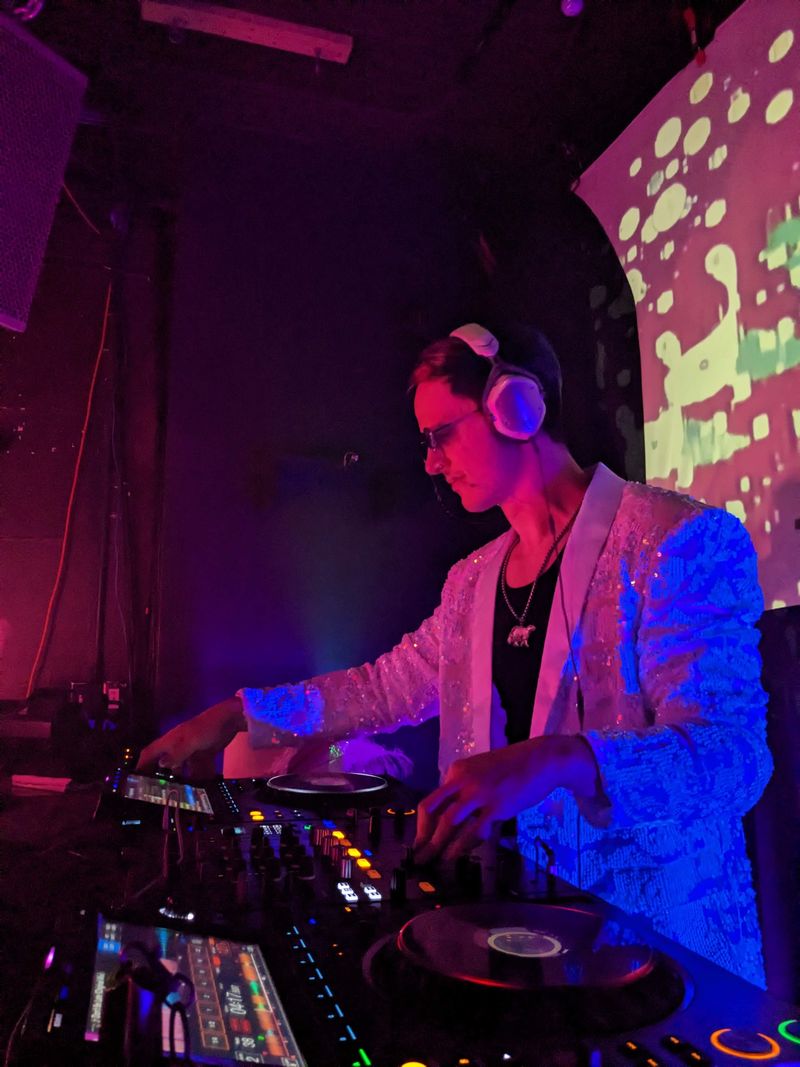chillbill in headphones and a white sequin jacket, working a Pioneer mixer under blue light